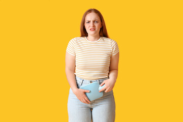 Young woman with hot water bottle having menstrual cramps on yellow background - obrazy, fototapety, plakaty