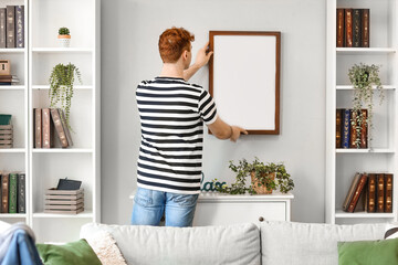 Young redhead man hanging blank frame on light wall at home, back view