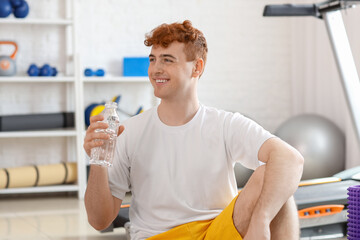 Young redhead man with bottle of water in gym