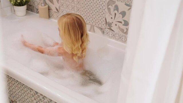 Laughing caucasian blonde three years old girl taking bath with white foam bubbles, playing, having fun, looking at camera.Carefree childhood, body washing, hygiene concept.