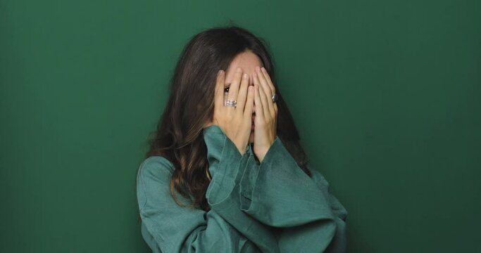 Scared shocked amazed young woman look camera covering hiding face with hands peep through fingers isolated on green background studio portrait. Shocked woman scary of movie.