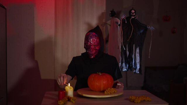 Person wearing glittery purple skeleton mask lights candles on Halloween table. Unrecognizable man sits at table with pumpkin on tray. Dark room is decorated with scary skeletons in robes.