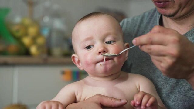 Baby feeding.Parent feed baby porridge.Child sits at table on chair with spoon. Happy kid eats porridge with spoon. Parent feeds the baby with a spoon. Father takes care of child. Family lunch at home