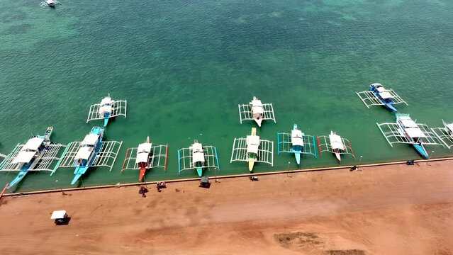 Aerial Drone Bird's Eye Top Down View of multiple Filipino Outrigger Bangka Boats moored to a quaint dock, revealing tropical village town and settlers inland. Vacation shot from the Philippines