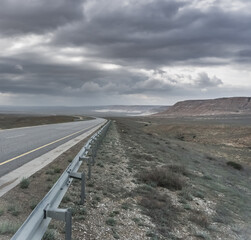 Asphalt road across the steppe and desert of Kazakhstan among the slopes of sandstone and lime, in cloudy weather with rain clouds