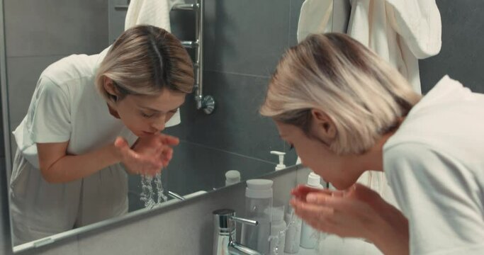 Happy attractive young fair-haired woman wash face remove makeup look at mirror, smile, enjoy natural beauty healthy clean soft moisturized skin care treatment close up view slow motion daily routine