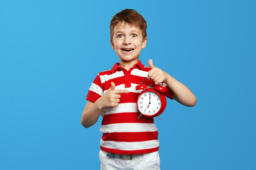 Stunned kid pointing with finger at classic bell alarm clock while looking at camera against blue