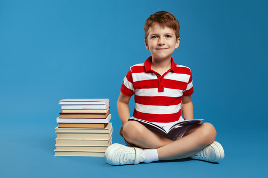 Cute kid holding open book while and looking at camera while sitting on floor near stack of books