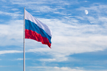 Large flag of the Russian Federation on a flagpole 75 meters high. The early moon hides behind the clouds in the background on a sunny evening. Blagoveshchensk, Far East, Russia