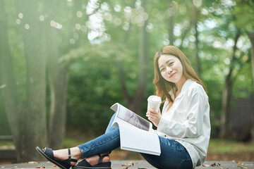 A young woman is reading a book while drinking coffee with a disposable paper cup in a forested park.