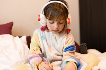 little girl in pajamas and headphones sits on the bed, draws with a pen in a notebook, horizontal format