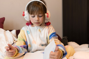 Fototapeta premium little girl in pajamas and headphones sits on the bed, draws with a pen in a notebook, horizontal format