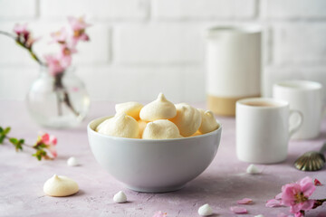 Small meringues in a white bowl over light pink background decorated with spring peach tree flowers, two cup of coffee behind - obrazy, fototapety, plakaty