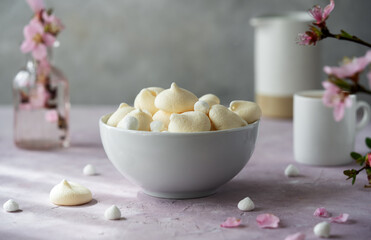 Small meringues in a white bowl over light pink and grey concrete background decorated with spring peach flowers, cup of coffee, creamer behind - obrazy, fototapety, plakaty