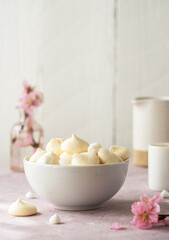 Small meringues in a white bowl over light pink and white wooden background decorated with spring peach flowers, cup of coffee and creamer behind