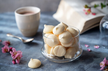 Small meringues in a glass bowl over grey concrete background decorated with spring peach flowers, cups of coffee, book behind - obrazy, fototapety, plakaty