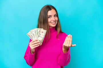 Young caucasian woman taking a lot of money isolated on blue background inviting to come with hand. Happy that you came