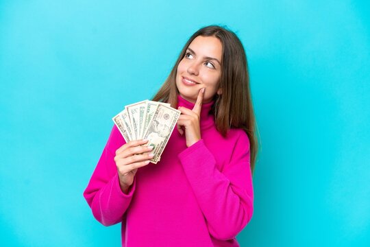 Young caucasian woman taking a lot of money isolated on blue background thinking an idea while looking up