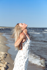 Fototapeta na wymiar Happy blonde woman in free happiness bliss on ocean beach standing straight. Portrait of a female model in white summer dress enjoying nature during travel holidays vacation outdoors