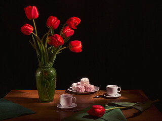 In a dark room, on a wooden table is a green vase with tulips. next to the table are two white cups and a plate of marshmallows