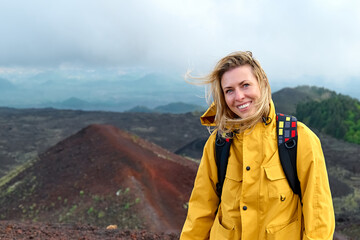 Hiking on tallest volcano in Continental Europe - Etna. Young smiling woman in yellow raincoat in...
