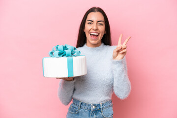 Young caucasian woman holding birthday cake isolated on pink background smiling and showing victory sign