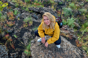 Hiking on tallest volcano in Continental Europe - Etna. Smiling young woman looking at camera near vegetation on black sand lava-rocky terrain of Mount Etna in background.