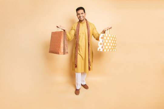 Happy young indian man wearing traditional kurta outfit holding shopping bags standing isolated on beige studio background. Diwali celebration and festive sale concept.