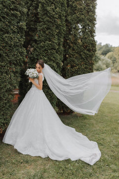 the bride in a wedding dress with a long train and a veil holds a wedding bouquet of roses against the background of tall green trees. Photo from the back