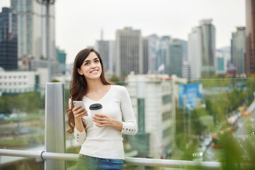 Fototapeta premium A young woman is resting on a land bridge with coffee and a smartphone in the background of a busy street during the day.
