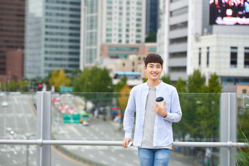 A young man is resting on a land bridge with coffee and a smartphone in the background of a busy street during the day.