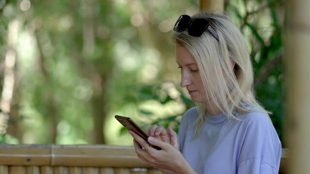 Young beautiful blonde woman sitting in park in gazebo and texting using mobile phone, smartphone. Sunny summer day, she works or relaxes in fresh air. Student.