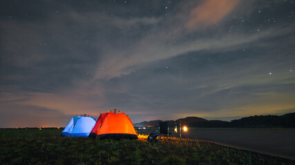 Fototapeta na wymiar riverside camping, blue and orange riverside tent camping with luggage, bonfire, evening camping activities The atmosphere of camping at night when the sky is full of stars.