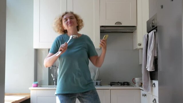 blond middle-aged woman dancing in kitchen with frying pan and ladle, emotion image, cooking love