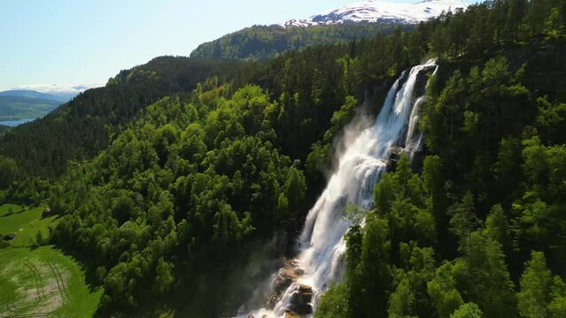 Drone footage around Tvindefossen waterfall with a green forested landscape on a sunny day