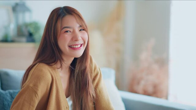 Confident smiling young adult asian woman looking at camera while sitting at home, Happy beautiful lady pretty face dental smile posing alone indoors, Slow motion close up view portrait