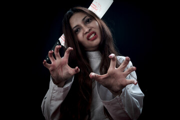 Beautiful black long hair woman with knife in head wearing white ghost spooky costume, standing with scary hand reaching out in dark background, going to celebrate Halloween party, play trick or treat