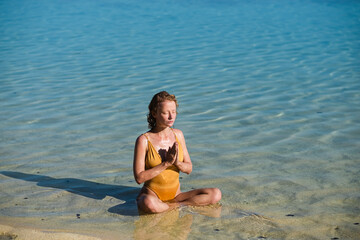 A young girl, with blond hair, of European appearance in a yellow swimsuit, practices yoga, and meditates on the beach with hands clasped in namaste.