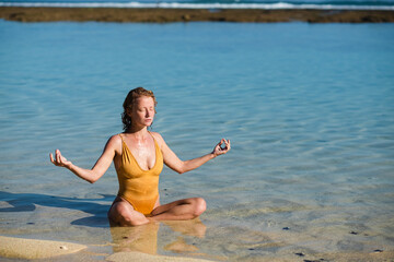 A young girl, with blond hair, of European appearance in a yellow swimsuit, practices yoga on the...