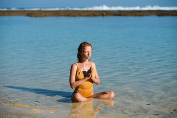 Fototapeta na wymiar A young girl, with blond hair, of European appearance in a yellow swimsuit, practices yoga, and meditates on the beach with hands clasped in namaste.