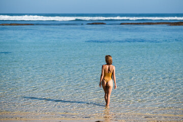 
A young girl in a yellow swimsuit, European appearance with blond hair, enjoys life on the beach and admires the ocean, rear view.