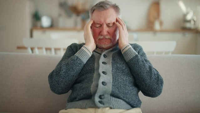 Senior man suffering from terrible headache touching head sitting on couch at home, making massage with closed eyes,doing turning head on sides exercise. Migraine, high intracranial pressure concept.