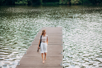 A beautiful girl with fashionable make-up and hairstyle, in white trousers and a beige top, with a black leather handbag, is walking along a boat pier on a picturesque river.