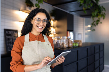 Portrait of a young beautiful woman at the workplace of a barista, a waitress, smiles and looks into the camera, an employee holds a tablet computer in her hands, works inside a cafe restaurant.