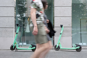 Shared electric kick scooters for rent in front of a fashion store window. A couple walks by (motion blur). E-mobility - obrazy, fototapety, plakaty