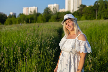 Copy space. Cute blond woman squinting stands in the summer in a green field in the rays of the setting sun. A squinty woman looks at the camera with one eye covered against a background of greenery.