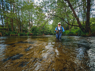 Man with fishing rod in green forest, standing by river, looking at camera