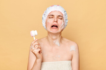 Crying unhappy woman wearing shower cap standing isolated over beige background doing beauty cosmetology procedures at home being in bad mood.