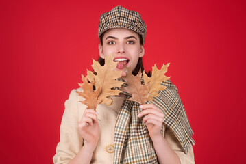 Obraz premium Hipster young woman in trendy autumn fall outfit stylish hat and scarf. Beauty girl hold autumn leaves near face, isolated on studio background. Portrait of autumnal woman with maple leaf.