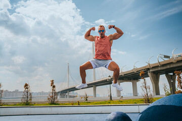 Muscular blond caucasian man in sportswear and sunglasses jumps high shows his biceps against view on big bridge and sky with clouds on summer sunny day. Fitness coach at outside training. - Powered by Adobe